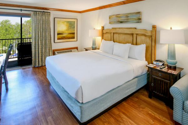 The image shows a hotel room with a king-sized bed, two nightstands with lamps, a chair, a desk, a TV, and a balcony with a view.