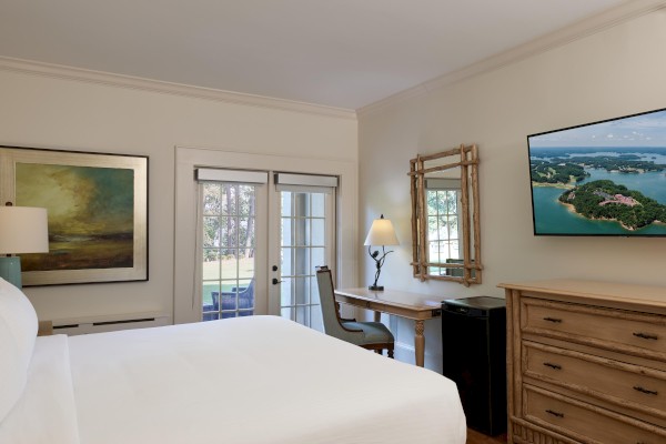 A cozy bedroom with a large bed, artwork, a TV, a desk with a lamp, and glass doors leading outside complete the scene.