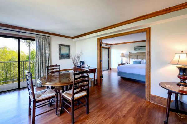 A spacious room features a dining table with chairs, a lamp on a side table, an open doorway to a bedroom with a bed, and a balcony with a scenic view.