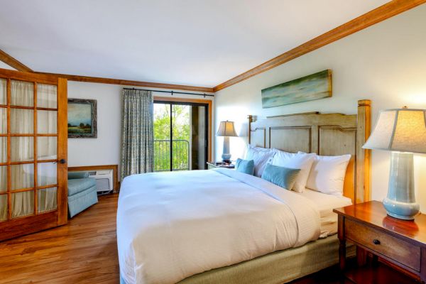 A cozy bedroom with a large bed, wooden headboard, bedside lamps, a small seating area, and a door opening to a balcony with a view.