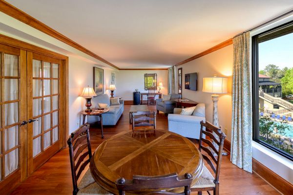 A spacious room with a dining table, chairs, a sitting area with sofas and armchairs, wooden flooring, French doors, and a large window with a pool view.