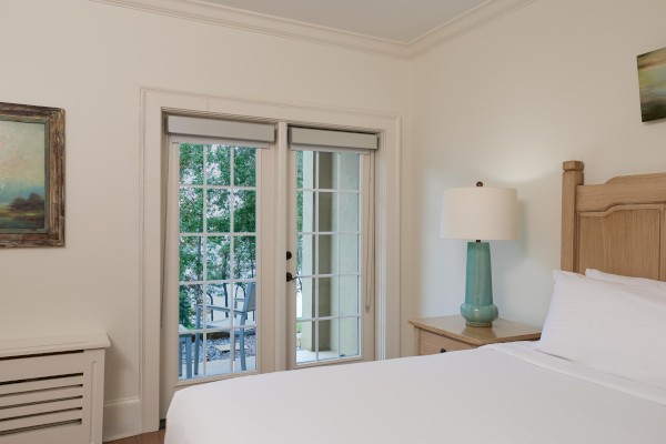 A bright bedroom with a bed, bedside lamp, and art on the walls. French doors open to a view of a balcony with chairs and greenery outside.