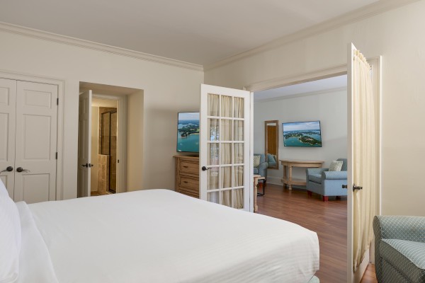 A hotel room with a bed, open French doors leading to a sitting area with armchairs, TV, and a wooden dresser.