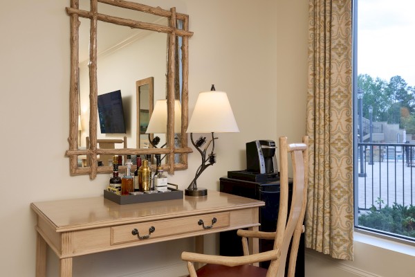 A wooden desk with a mirror, lamp, coffee maker, and assorted bottles next to a window with curtains and a view outside.