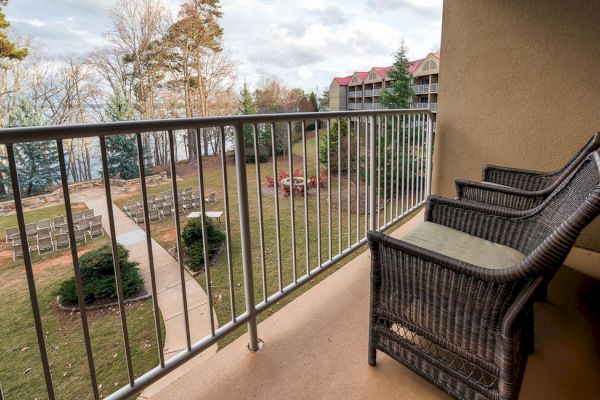 A balcony with wicker chairs overlooks a landscaped garden with a walkway, trees, and a distant building visible in the background.