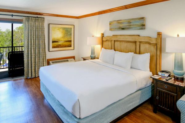 This image shows a neatly arranged hotel room with a large bed, bedside tables, lamps, an armchair, art on the walls, and a balcony with a scenic view.