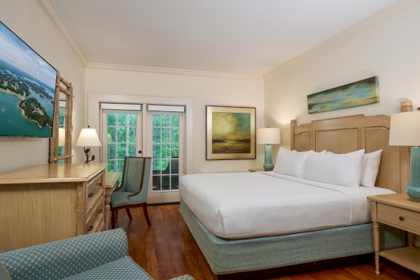 A cozy bedroom with a large bed, wooden furniture, wall art, a TV, and a view of greenery through glass doors.