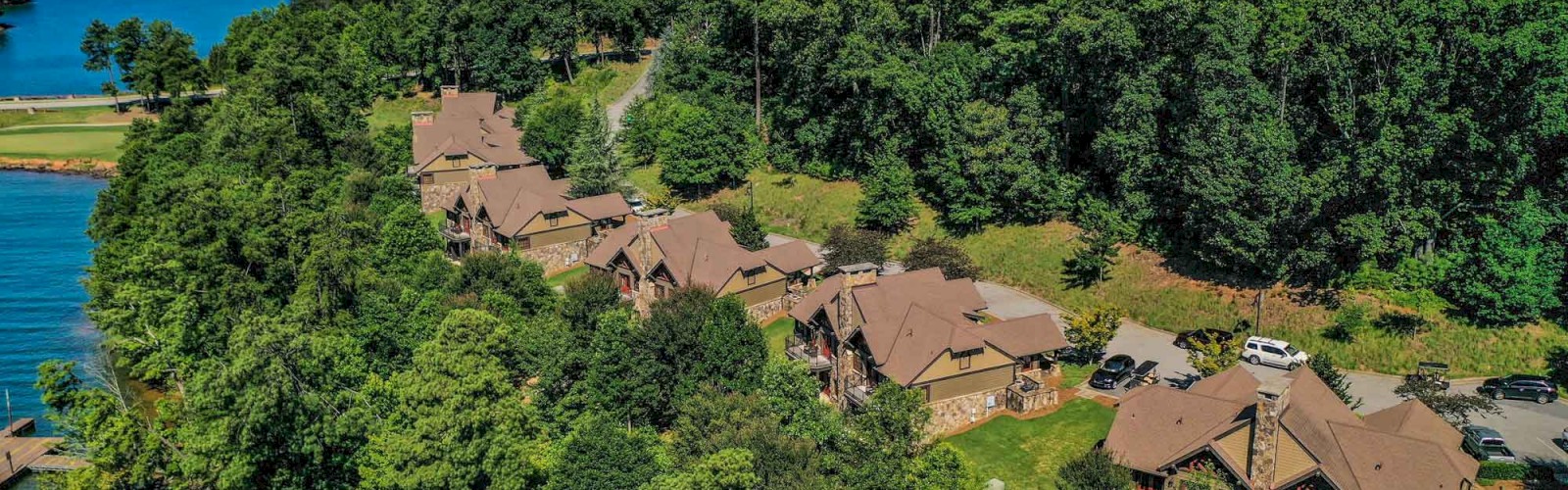 The image shows a row of cabins nestled among trees alongside a lake, with a backdrop of forested hills and a clear sky above.
