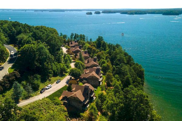 Aerial view of houses nestled among trees near a vast body of water with scattered islands and boats in the distance, along a curving road.