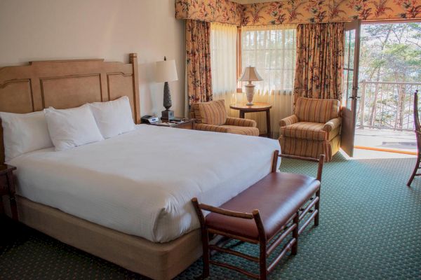 The image shows a cozy hotel room with a large bed, bedside tables, two chairs, a lamp, and a small table by the window. A balcony is also visible.