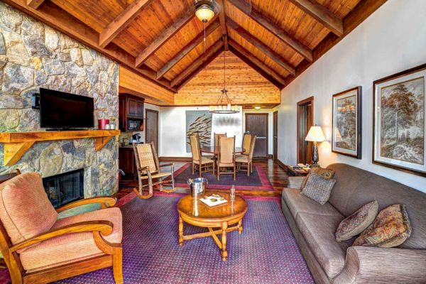 A cozy living room features a stone fireplace, TV, wooden furniture, brown sofa, armchair, round table with champagne, leading to a dining area.
