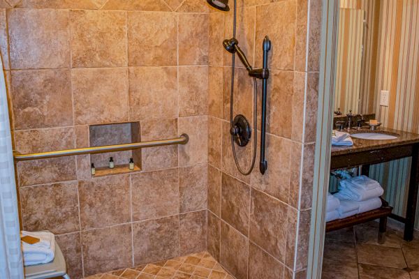 A tiled shower with a grab bar, showerhead, and niche, adjacent to a vanity with towels and toiletries on a wooden shelf.