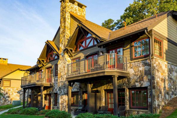 The image shows a large, rustic-style house with stone façades, wooden balconies, and surrounded by greenery and trees.