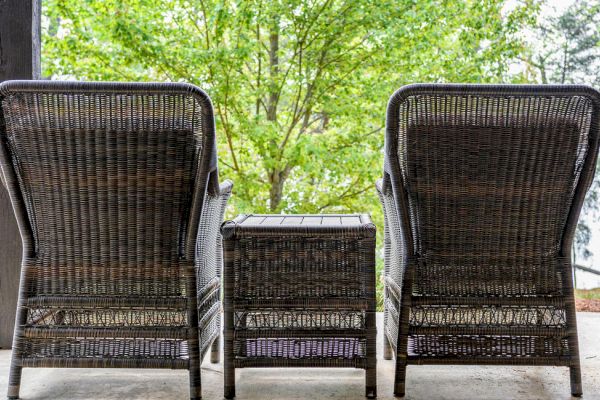 Two wicker chairs with a small wicker table between them, placed on a patio with a green, leafy tree and scenic view in the background.