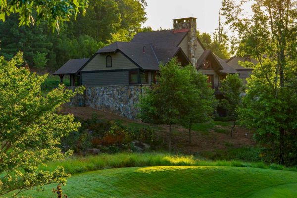 A stone and wood cabin nestled in a forested area, surrounded by lush greenery and trees, with sunlight filtering through the foliage.