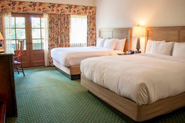 A cozy hotel room with two double beds, a desk with a lamp, a dresser, a chair, and a balcony door. The room is brightly lit and inviting.