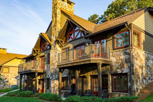 The image shows a beautiful building with a stone exterior and wooden accents, featuring balconies and surrounded by greenery, under a clear sky.