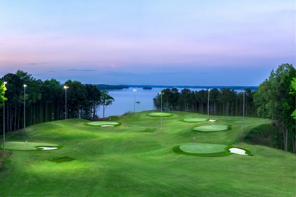 A scenic golf course by a lake, with multiple greens, sand traps, trees, and bright lights under a twilight sky.