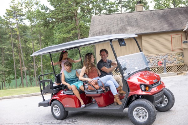A family of four enjoys a ride in a red golf cart near a house with a stone wall and trees in the background.
