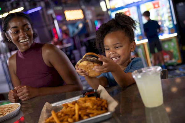 A woman and a girl are enjoying some food at a restaurant, with the girl holding a large burger, smiling excitedly.