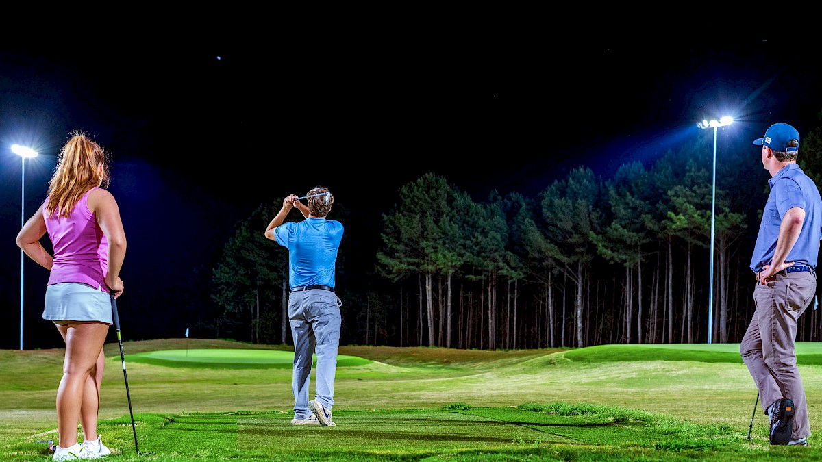 Three people play nighttime golf on a well-lit course with a backdrop of trees.