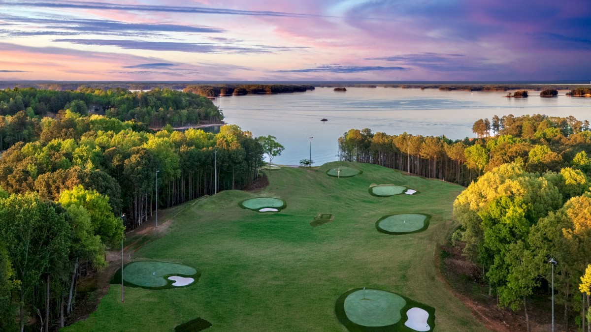 A scenic view of a golf course near a large body of water during sunset, with trees, putting greens, and sand bunkers visible.