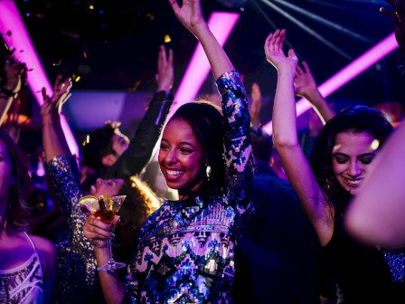 People dancing at a nightclub, smiling and raising their hands; colorful lights, party vibes, and a woman in the center holding a drink.