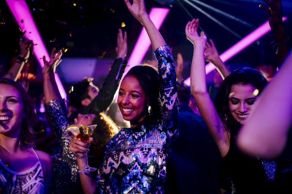 People dancing at a nightclub, smiling and raising their hands; colorful lights, party vibes, and a woman in the center holding a drink.