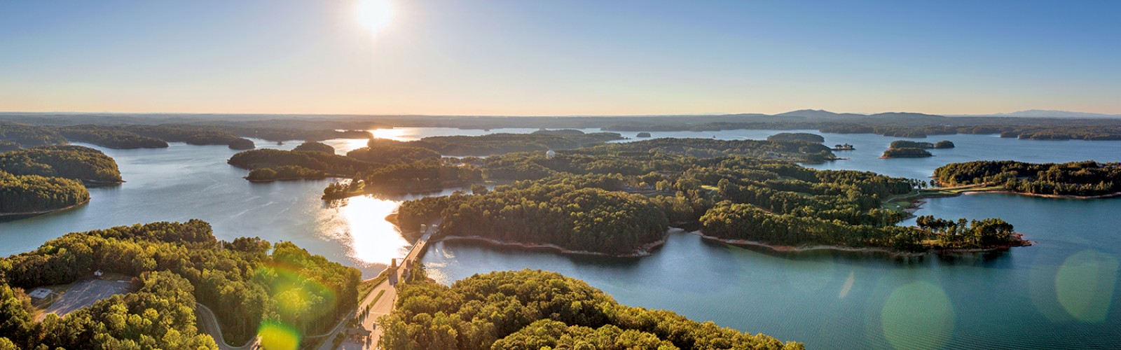 Aerial view of a lake with numerous islands, surrounded by green trees, with the sun shining brightly in a clear sky.