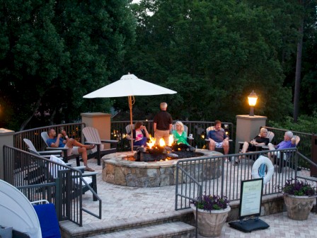 People are sitting around a lit outdoor fire pit on a patio, enjoying evening outdoors under trees and street lamps.