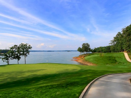 A scenic golf course by a lake with a winding path, green grass, trees, and a blue sky with wispy clouds.