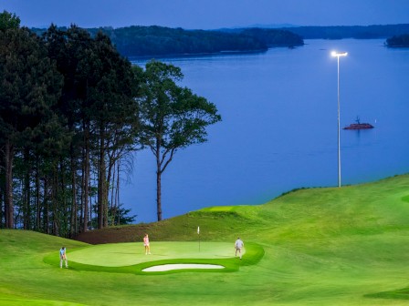 Golfers are playing under lights on a lush green course near a large body of water at dusk.