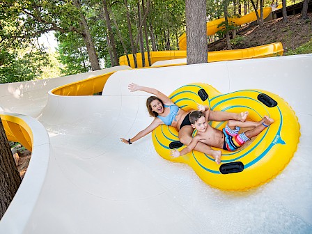 Two kids and an adult enjoy slipping down a white water slide at a forested park on a sunny day.