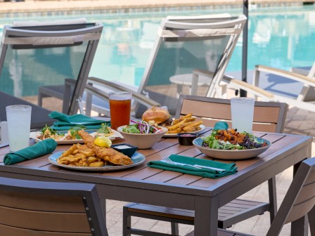 A poolside dining table with plates of fish and chips, burgers, salads, drinks, and napkins, near lounge chairs by a swimming pool.