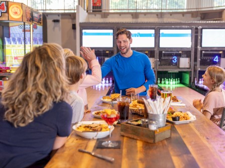 A family enjoys a meal together at an arcade, with bowling lanes visible in the background and everyone smiling and high-fiving.