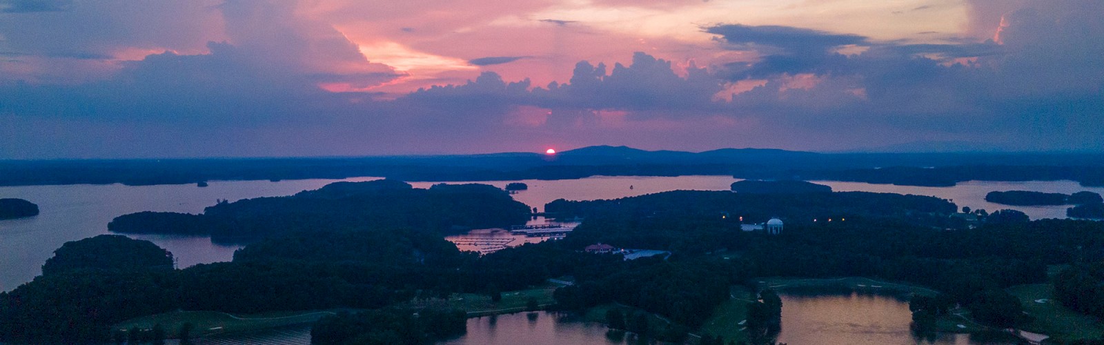 A serene lake landscape at sunset, with scattered islands, calm waters, and a sky painted in hues of pink, purple, and orange, seen from an aerial view.