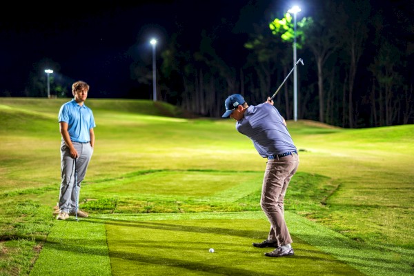 Two men are on a golf course at night, one preparing to hit the ball while the other watches under bright lights.