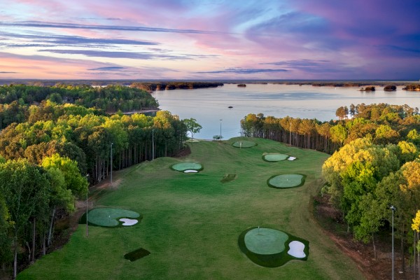 A scenic view of a golf course near a lake during sunset, with green fairways, trees, and a colorful sky.