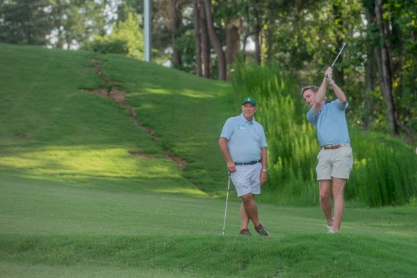 Two men are on a golf course, one is swinging a golf club while the other watches and smiles in the background.