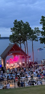 An outdoor concert at dusk near a lake with boats, trees, and an audience enjoying live music on a stage with colorful lighting.