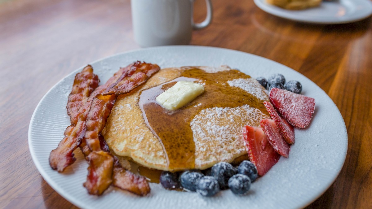 A plate of pancakes with butter, bacon, strawberries, blueberries, powdered sugar, and syrup, along with a cup of coffee in the background.