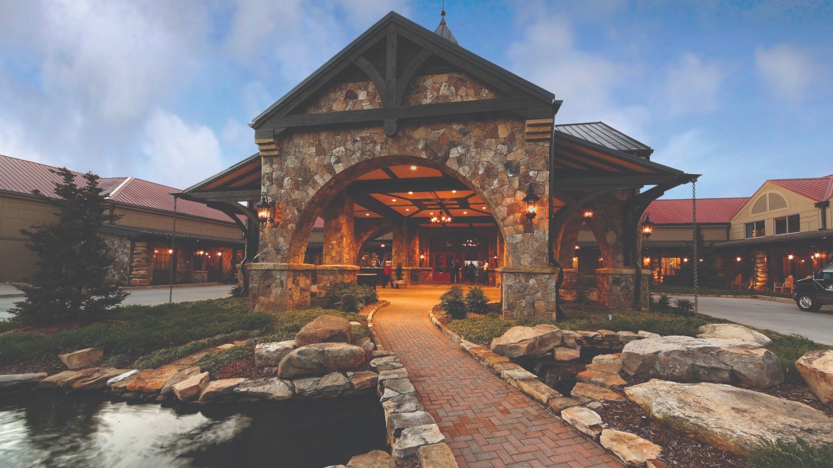 A charming stone and wood lodge with a welcoming entrance, surrounded by landscaped gardens and a small pond at dusk.