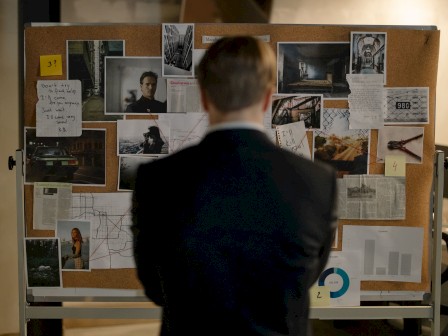 A man in a suit stands with his back to the camera, observing a corkboard full of photos, notes, and maps, possibly planning an infiltration.