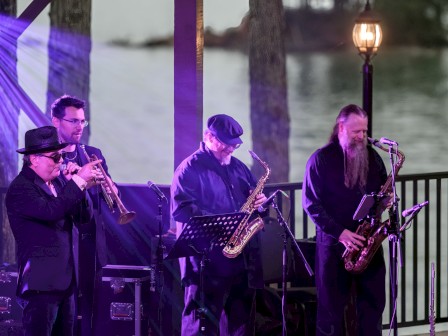 A live jazz band featuring four men playing instruments outdoors near a river at sunset with a vintage lamp post.