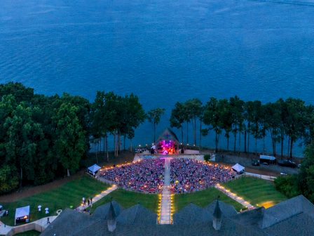 A nighttime outdoor concert takes place by a lake, with a colorful sky, surrounded by trees and a building at the bottom of the image.