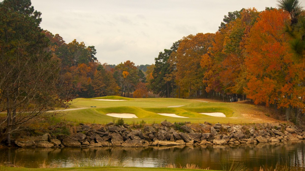 A golf course with sand bunkers, surrounded by colorful autumn trees and a calm water body in the foreground.