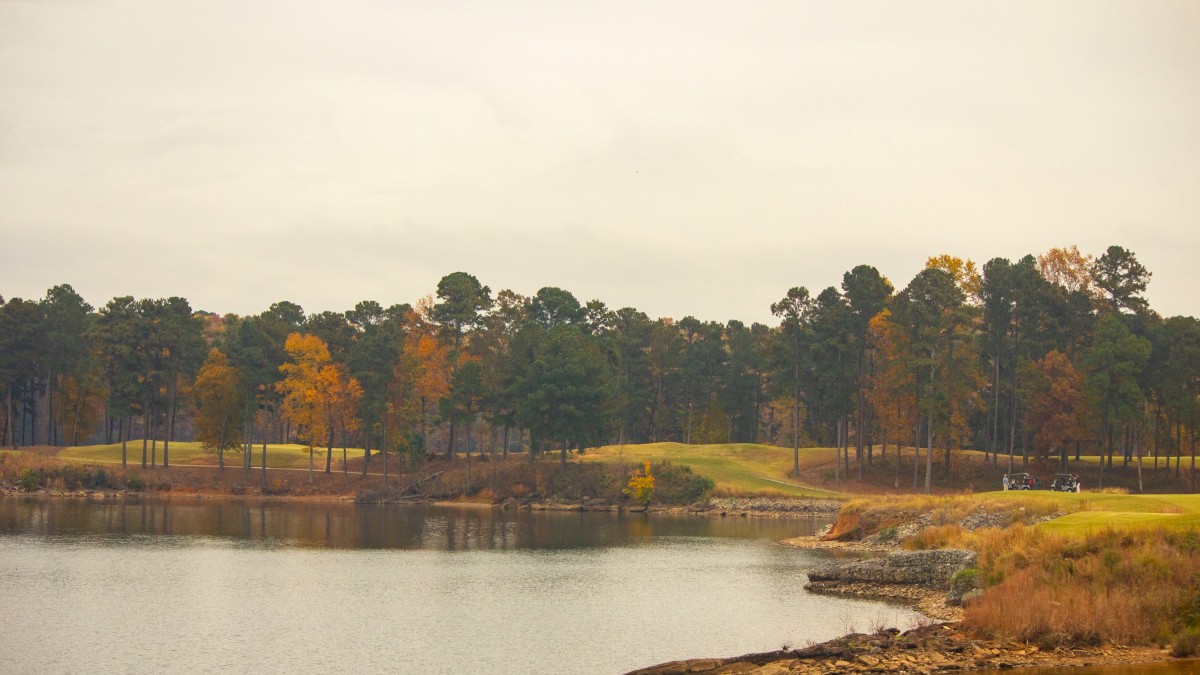 A peaceful lake scene with a forested shoreline, some boats, and autumn-colored trees under a cloudy sky.