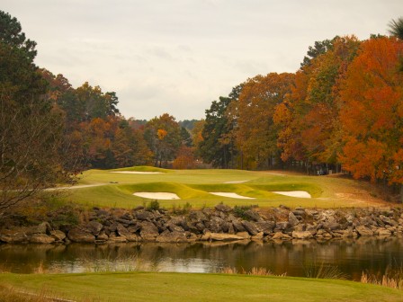 A scenic golf course with sand bunkers, surrounded by autumn-colored trees and a calm water body reflecting the landscape.