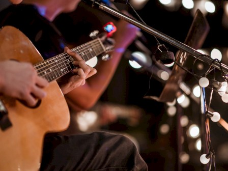 A person is playing an acoustic guitar during a live performance, with string lights and a microphone in a cozy, dimly lit setting.