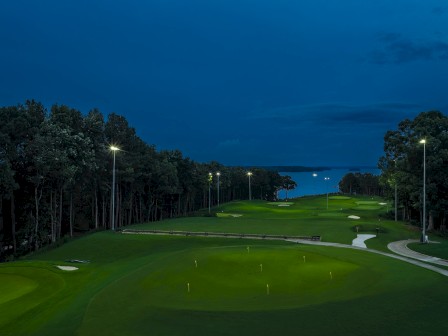 A nighttime view of a golf course with lit lampposts, green grass, trees, and a distant water body under a dark cloudy sky.